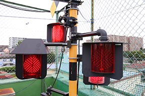 踏切警報機柱（アルミ形）に装着された、踏切警標と全方向踏切警報灯 LED III形。そのうち1つ（左）はオプションの防雪フード付き