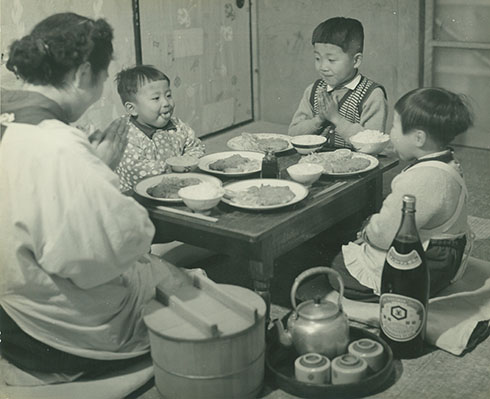 食卓を囲む家族　手前にラベルが見えるようにしょうゆ瓶が置かれている　母と兄がいただきますポーズ