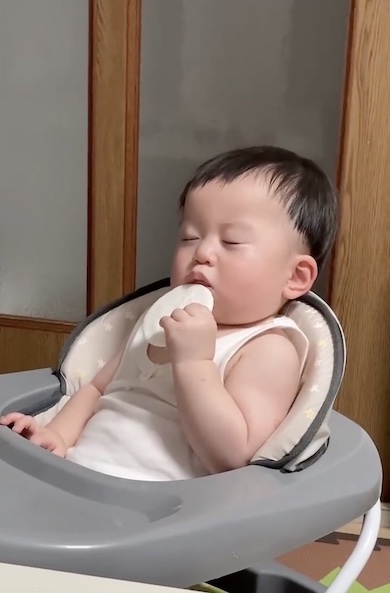 眠いまま口におせんべいを運ぶ赤ちゃんの写真