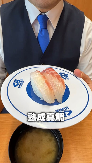 くら寿司メニューの1つ「熟成真鯛」を手に取っているところ。テロップに「熟成真鯛」と書かれている