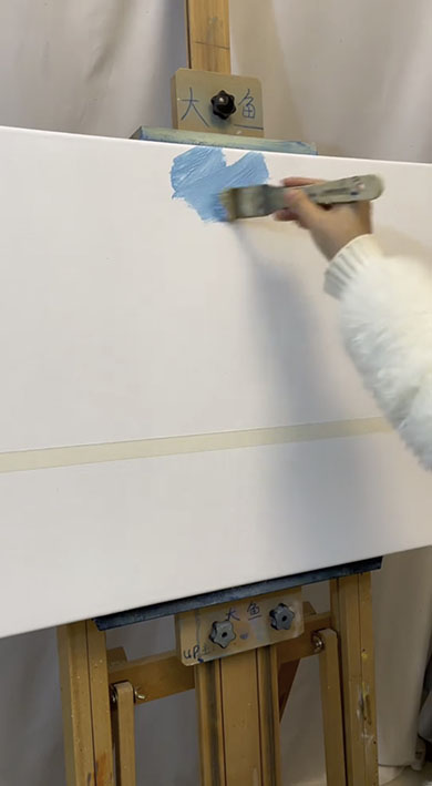白いキャンパスにブラシで青い絵の具を塗っているところ。真ん中上部に色を乗せている