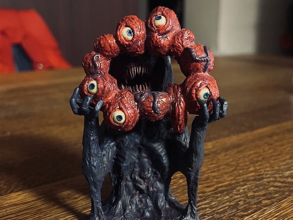 ミャクミャク風フィギュアの正面からの写真。赤く膨れた複数の目玉でできた輪の奥に、ギザギザの鋭い歯を持つ口が見える。