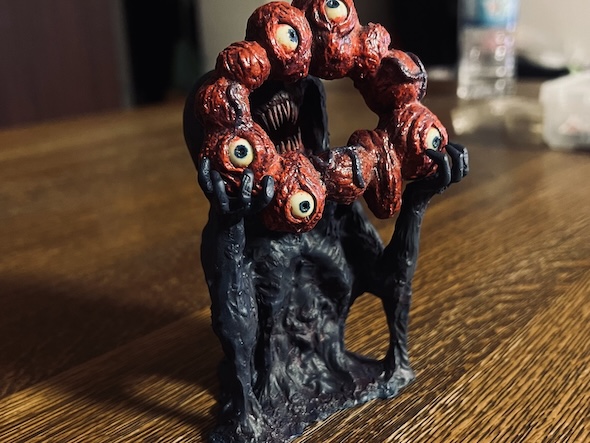 ミャクミャクをモチーフにした、ホラー調のフィギュア。黒くうねる体から赤い目玉の輪を掲げ、リアルな質感で異形の存在感を強調している。