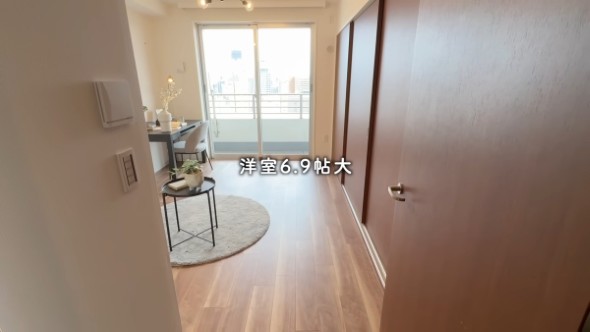 洋室6.9帖大と表示された、バルコニーに面した明るい洋室。木目調のフローリング、グレーのテーブルとチェア、小さな観葉植物が置かれた丸いラグの上のサイドテーブルが見える