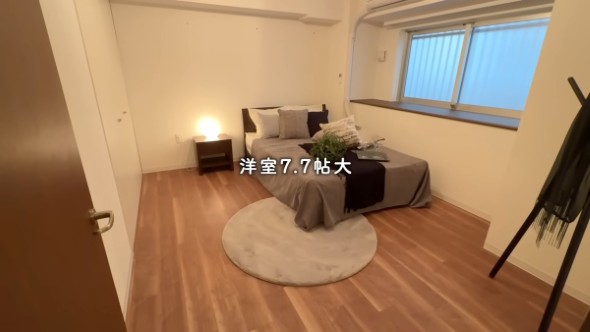 ベッドとサイドテーブル、コートハンガーが置かれた洋室が写っている。「洋室7.7帖大」とテキストが書かれている。