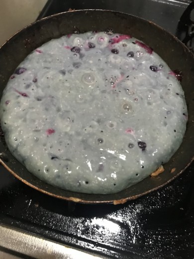 ブルーベリー入りのホットケーキ生地をフライパンで焼いている途中の様子。生地は灰紫色で泡が立っており、ところどころにブルーベリーが浮かんでいる。