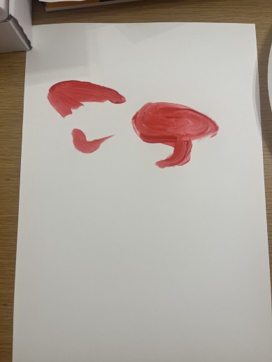 白い画用紙に赤い絵の具で筆跡が3つ描かれており、左上には髪型のような形、中央下に口元のような形、右側にはもう一つの髪型のような形がある。