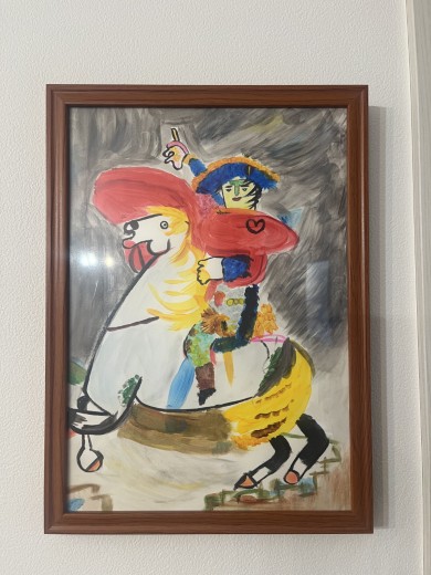 額縁に入れられたカラフルな絵画。赤いたてがみと黄色いしっぽの馬に、帽子をかぶった人物がまたがっている。人物は緑の顔をしており、服は青やピンク、黄色など多彩な色で塗られている。