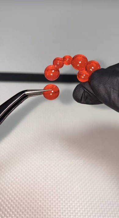 黒い手袋をした手とピンセットで赤い球状のパーツを組み立てている様子。複数の赤い球体が弧を描くように並んでおり、背景には『@candyman808』のウォーターマークが薄く表示されている。