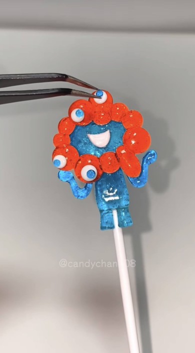 赤い球状の飴で囲まれた青い棒付きキャンディがピンセットで調整されている様子。キャンディーは複数の目と笑顔が描かれたキャラクターの形をしており、背景には『@candyman808』のウォーターマークが表示されている。