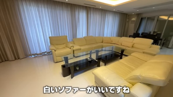 白いレザーソファがコの字型に並ぶ広々としたリビングダイニング。ガラスのローテーブルが中央にあり、背景には長いカーテンが広がっている。「白いソファーがいいですね」という字幕付き。