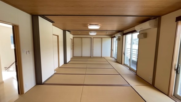 襖と障子に囲まれた広い和室。畳が敷き詰められ、複数の引き戸とエアコンが見える。窓の外にはバルコニーと景色が広がっている。