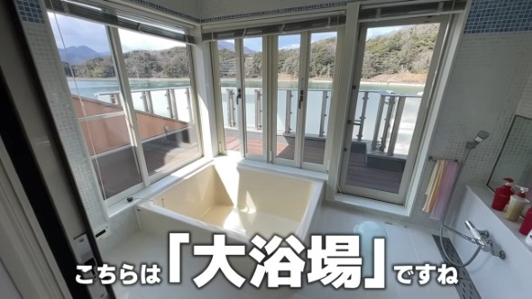 ガラス張りの窓に囲まれたバスルームで、大きな浴槽とシャワーエリアがある。外にはウッドデッキが見える。「こちらは『大浴場』ですね」というテキストが画面下部にある。