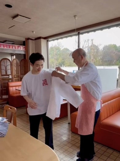レストランの店内で、年配の料理人が笑顔で若い男性に白衣を手渡すシーン。若い男性は白いTシャツを着ており、嬉しそうにそれを受け取っている。