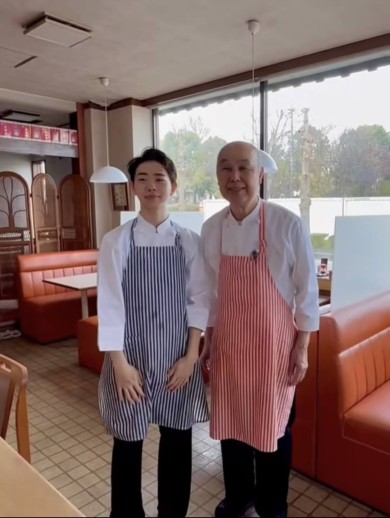 レストランの店内で、ストライプ柄のエプロンを着た若い男性と年配の男性が並んで笑顔で立っている様子。2人は祖父と孫の関係。