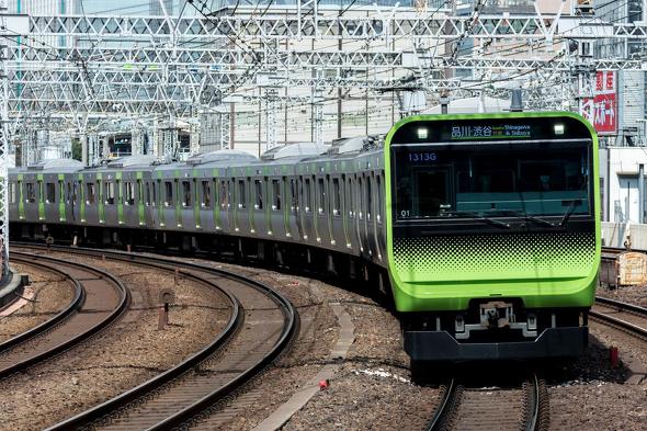 カーブした線路を走行する、東京の山手線の最新型車両。ライトグリーンのフロントデザインが特徴で、「品川・渋谷」行きの表示が見える。背景には複雑に組まれた架線やビル群が立ち並ぶ都市の風景が広がっている。